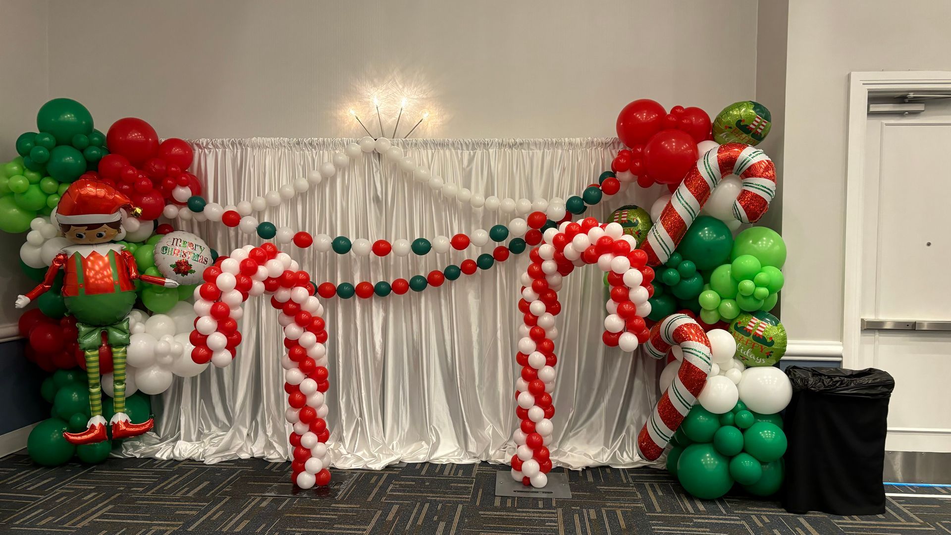Christmas-themed balloon arch with red, white, green balloons, candy canes, and an elf figure in front of a draped backdrop.