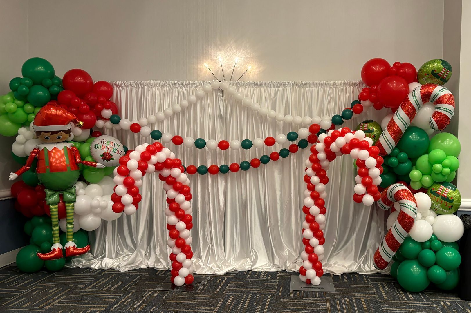 Christmas balloon display: elf, candy canes, green, red, and white balloons, draped white backdrop.