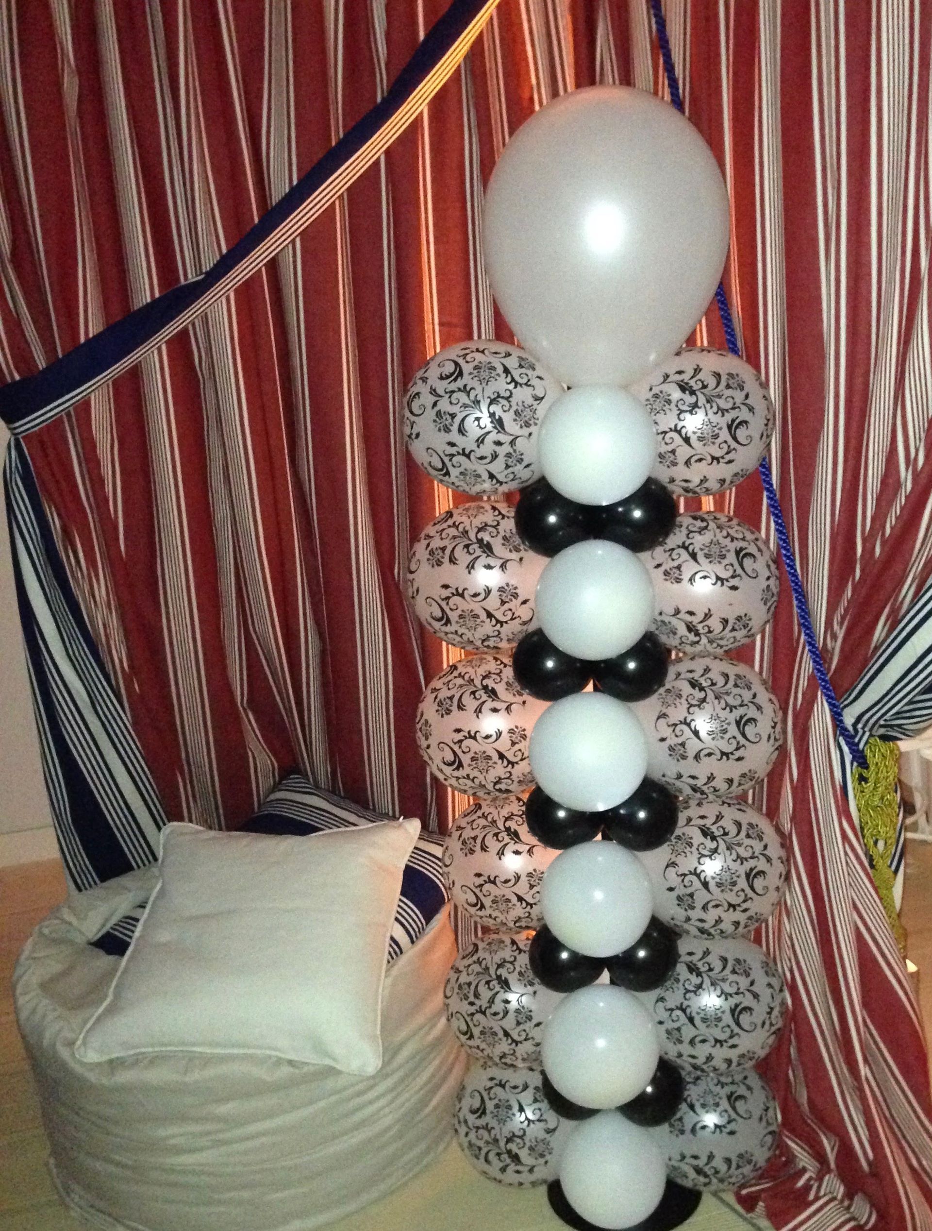 Balloon column with white, black, and confetti-print balloons; a white pillow on a round pouf is to the left.
