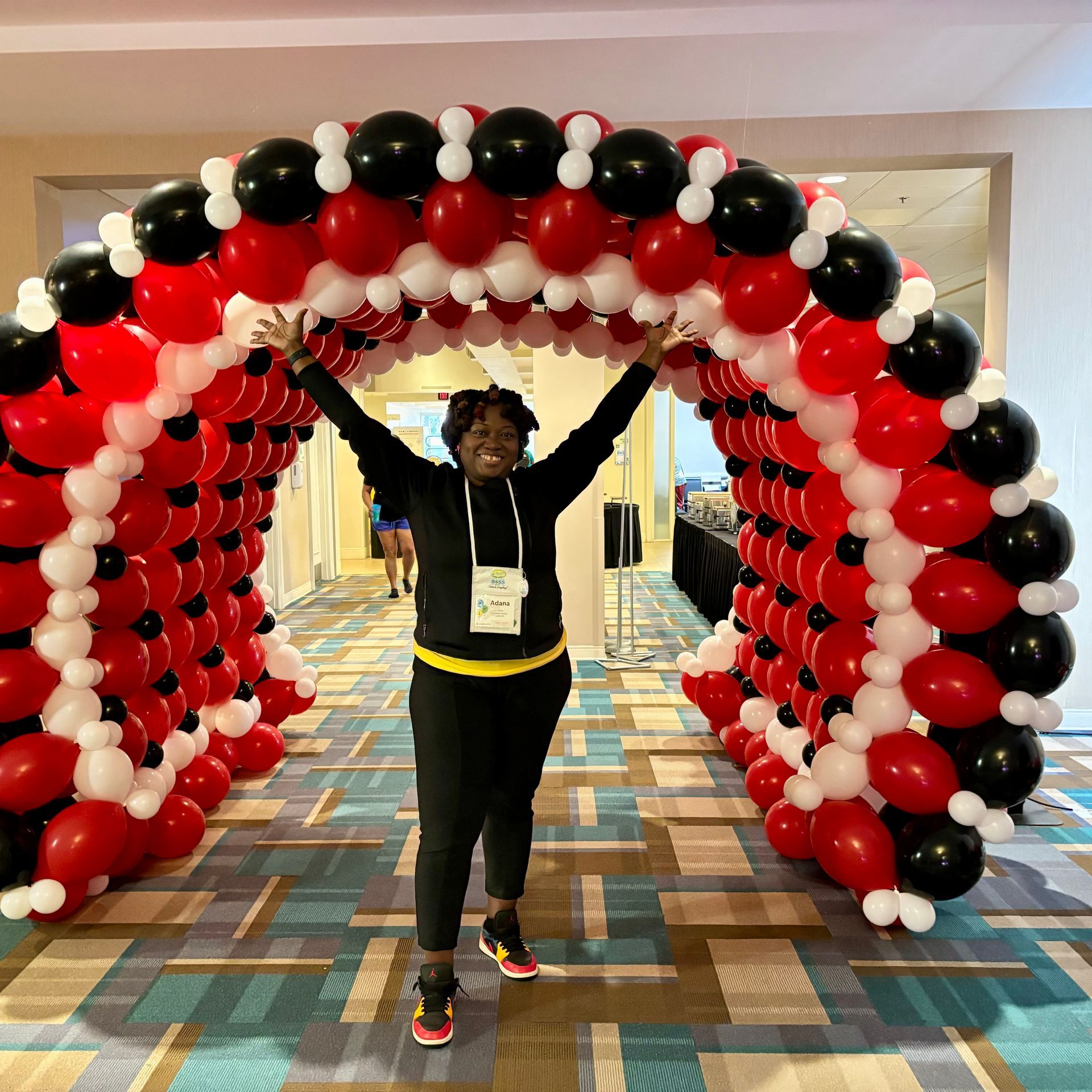 Woman with arms raised under a red, black, and white balloon arch. She is in a hallway.