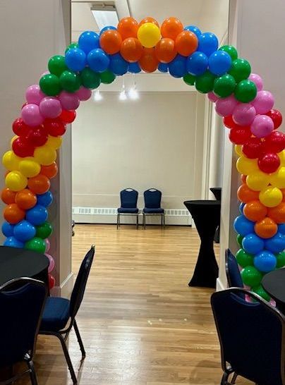 Rainbow-colored balloon archway frames an opening. Tables and chairs in a room.