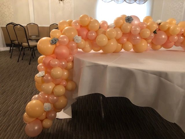 Balloon garland arching over a white tablecloth; orange and pink balloons, floral accents, indoor setting.