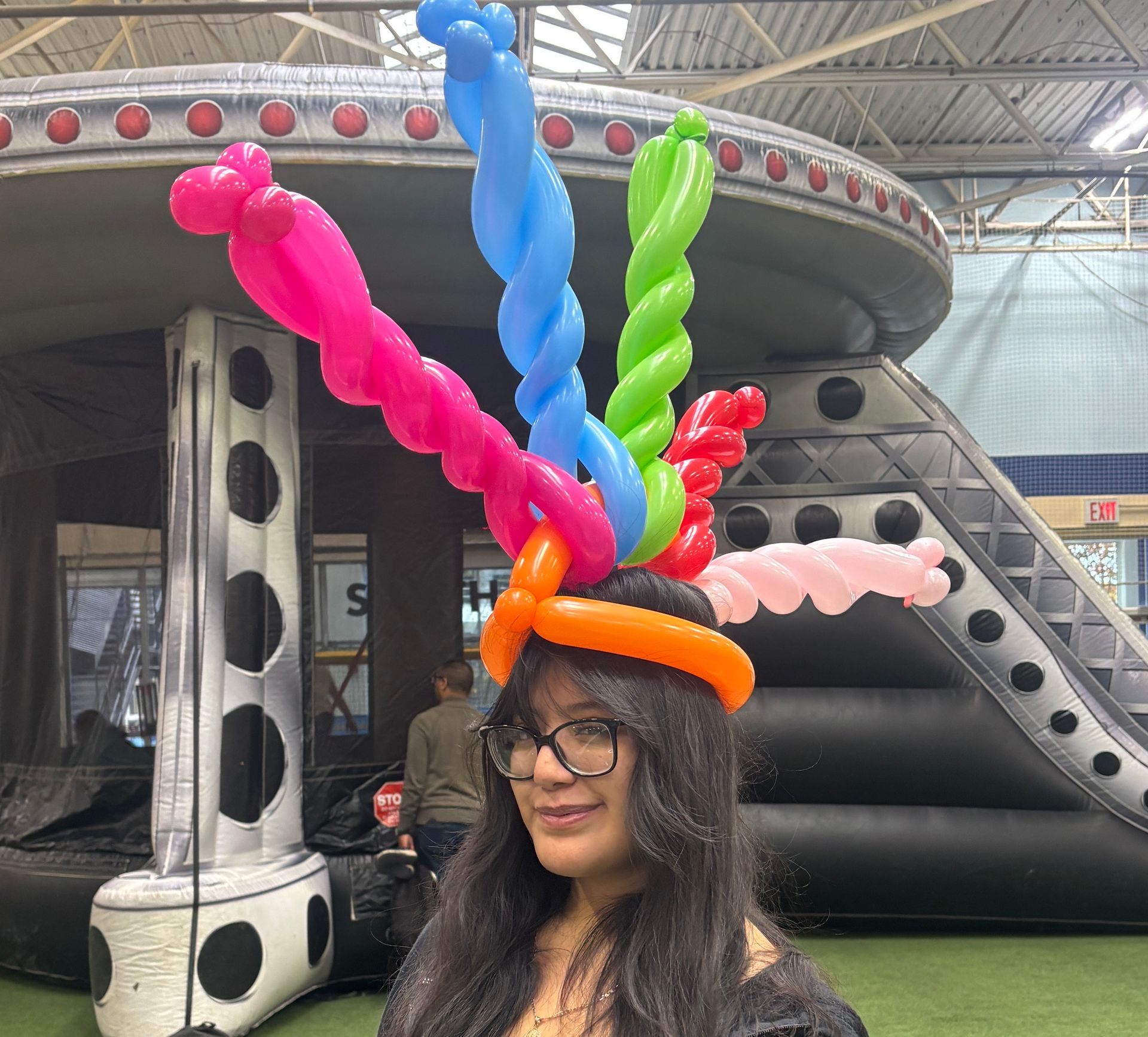 Woman wearing a colorful balloon hat smiles, standing in front of an inflatable structure with a slide at Chelsea Piers Field House Private event.