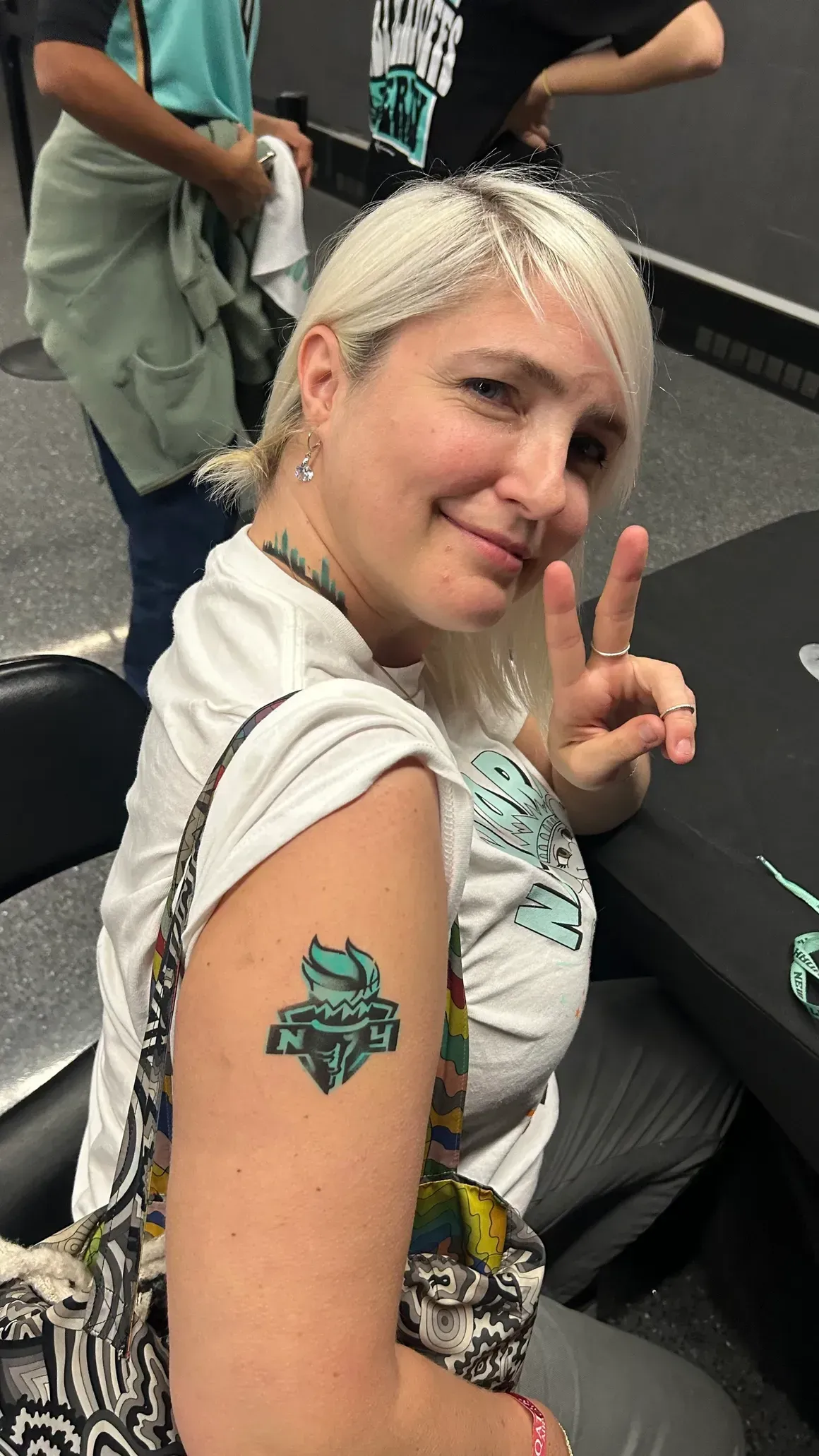 Woman with arm tattoo, giving peace sign. She is smiling, indoors.
