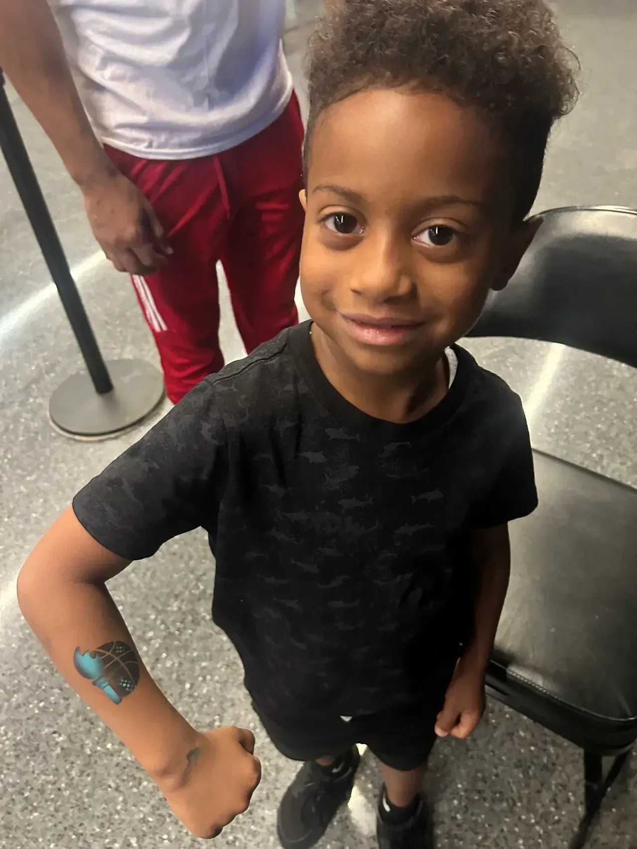 Boy with a temporary tattoo smiles, standing near a chair, wearing a black shirt and shorts.