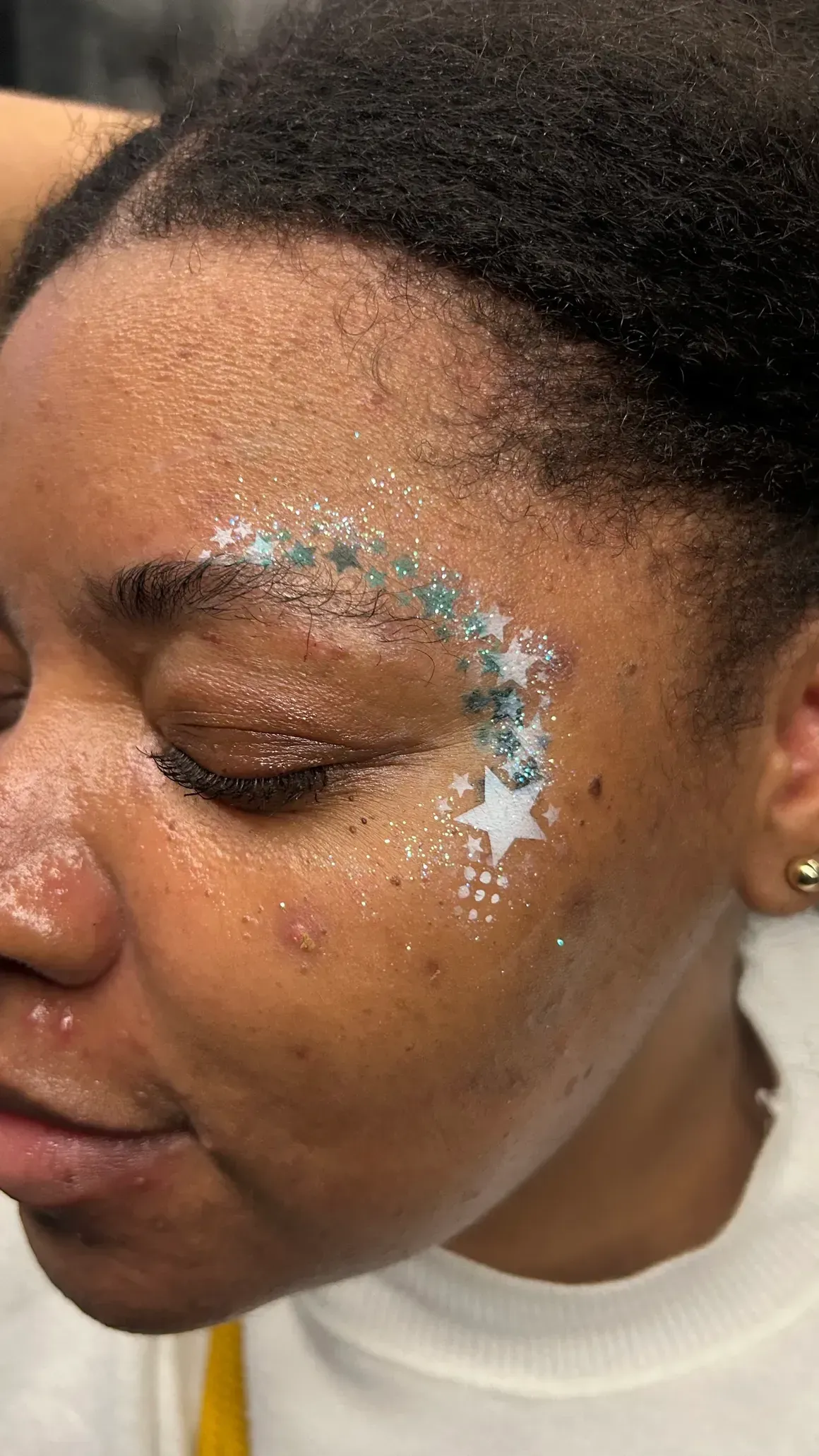 A person with dark skin has glitter makeup on their face in a starburst pattern.