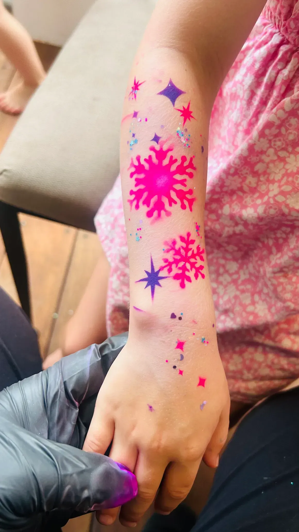 Child's arm with temporary tattoo of pink snowflakes and stars, being held by person with black gloves.
