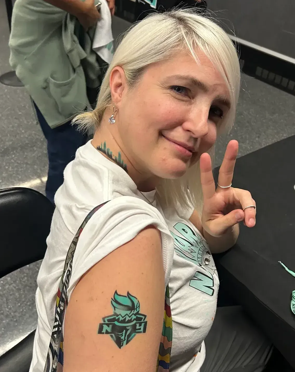 Woman with platinum blonde hair smiles, holding up peace sign. She has a teal tattoo on her arm.