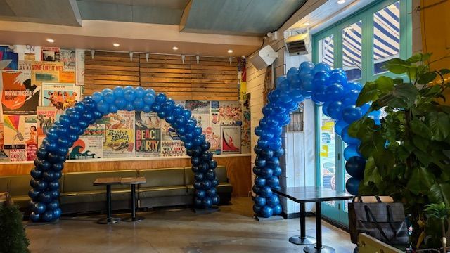 Two blue balloon arches in a cafe. The left arch is ombre. Tables are in the foreground, posters on the wall.