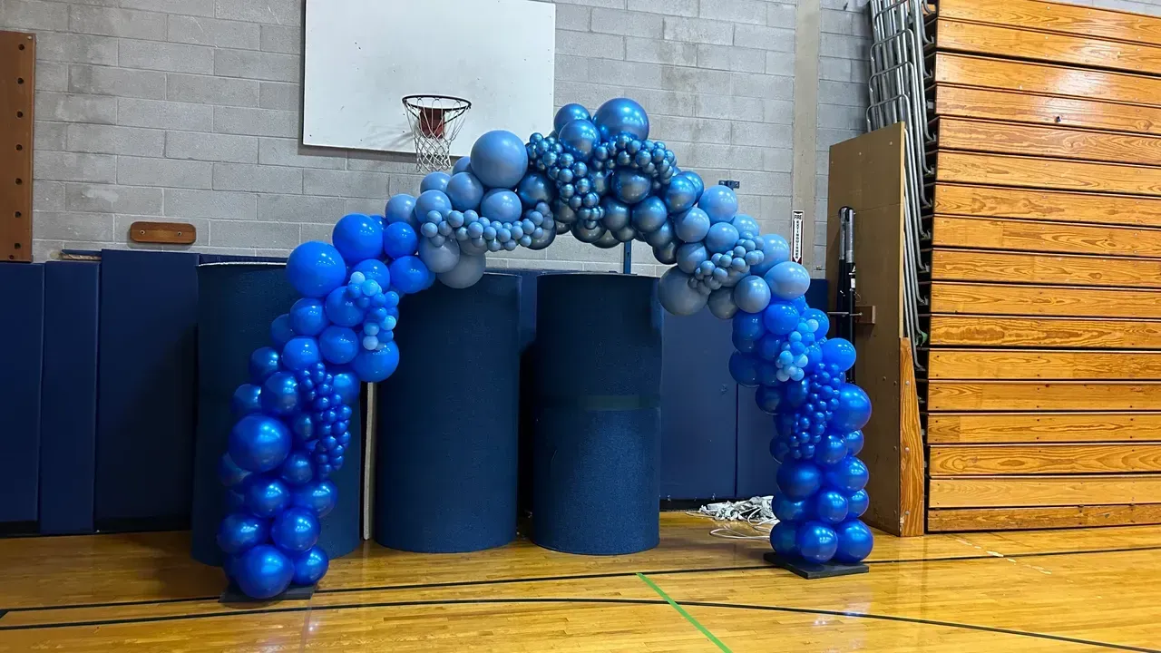 A blue and silver balloon arch in a gym with a basketball hoop in the background.