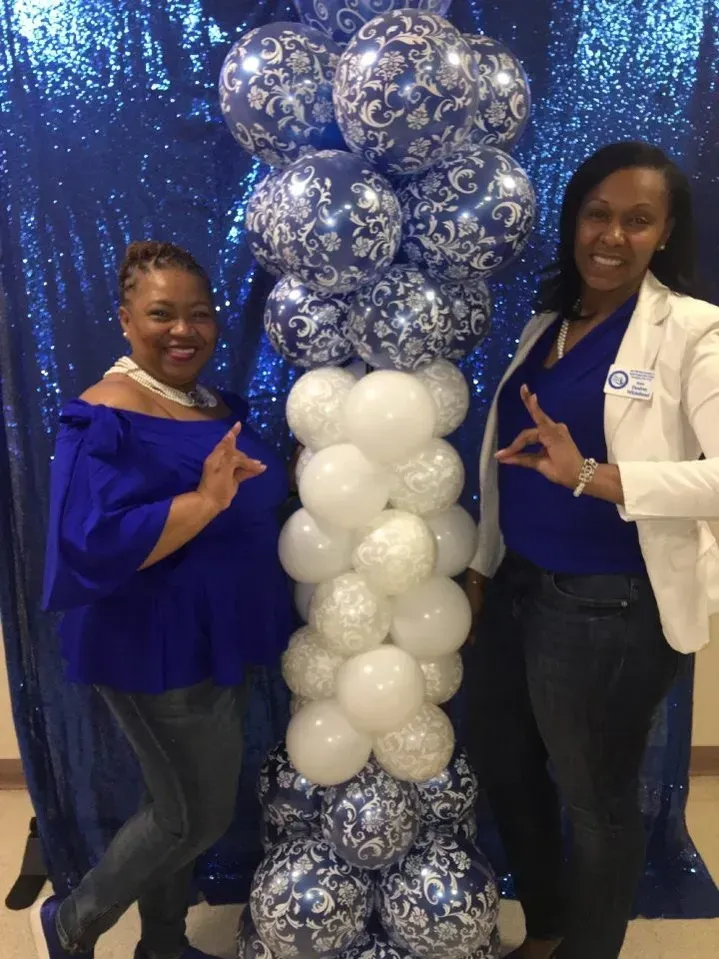 Two people posing by a blue and white balloon column and backdrop. Both wear blue and make Greek letter hand signs.