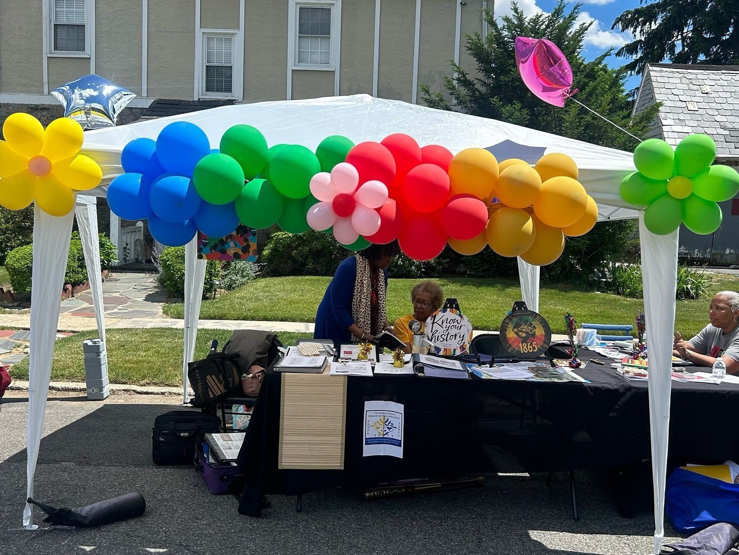 Booth with rainbow balloons, displaying items. People are seated inside. Bright, outdoor setting.