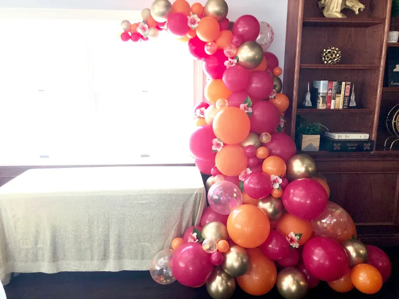 Balloon arch in red, orange, and gold, cascading over a table next to a window and bookshelf.
