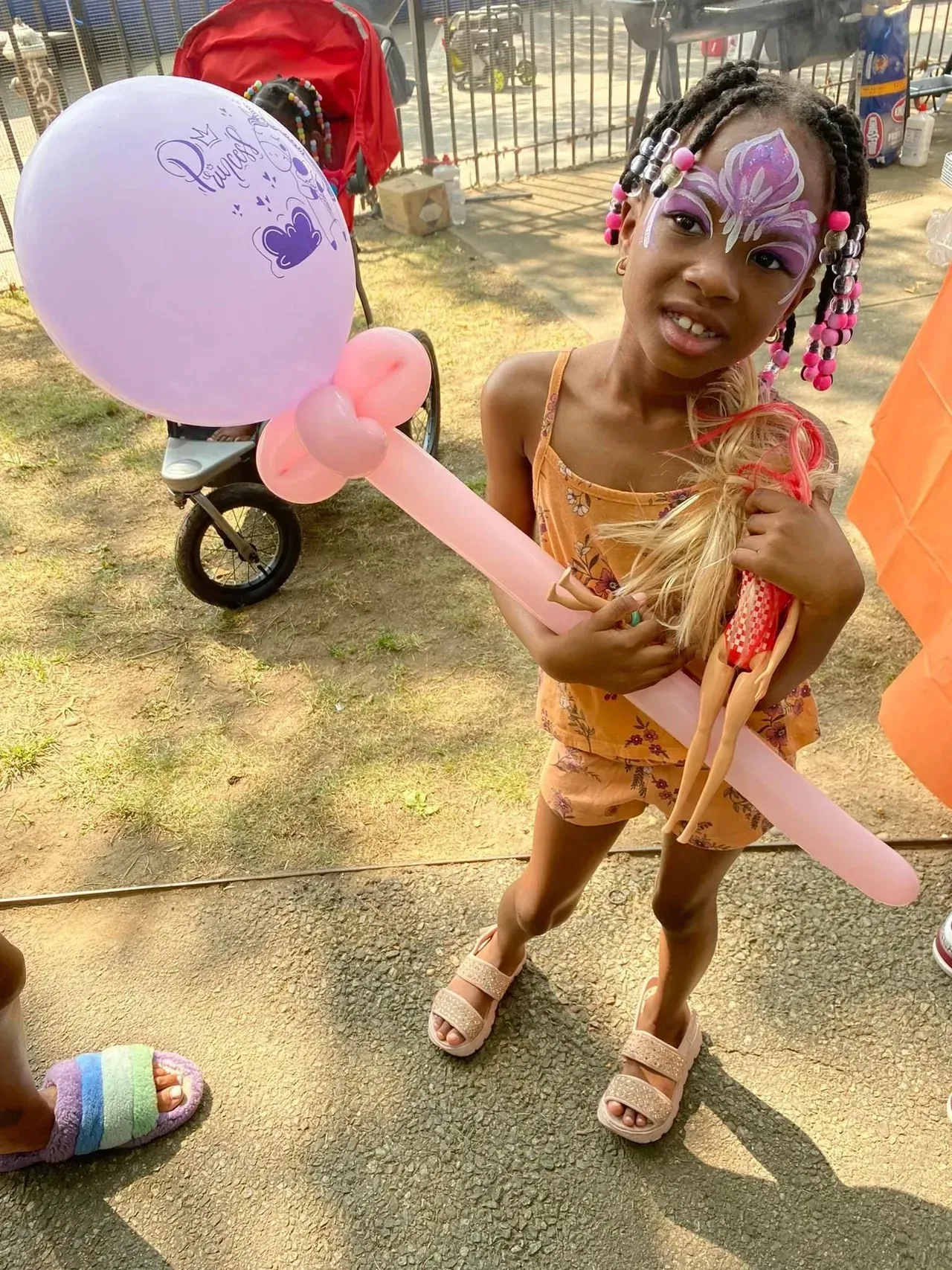 Girl with face paint holding balloon and doll, outside on a sunny day.