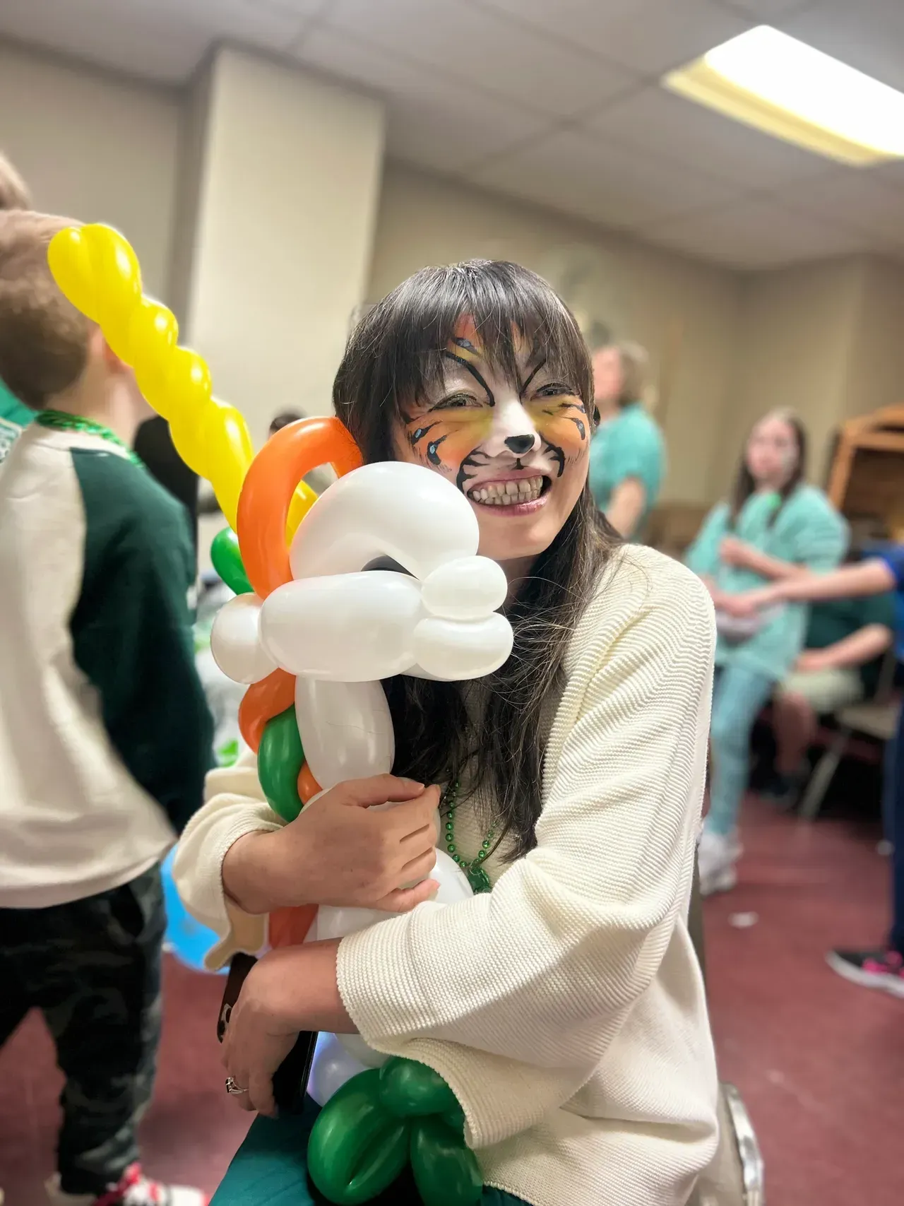 A woman with balloons painted on her face is holding a balloon shaped like a tiger.