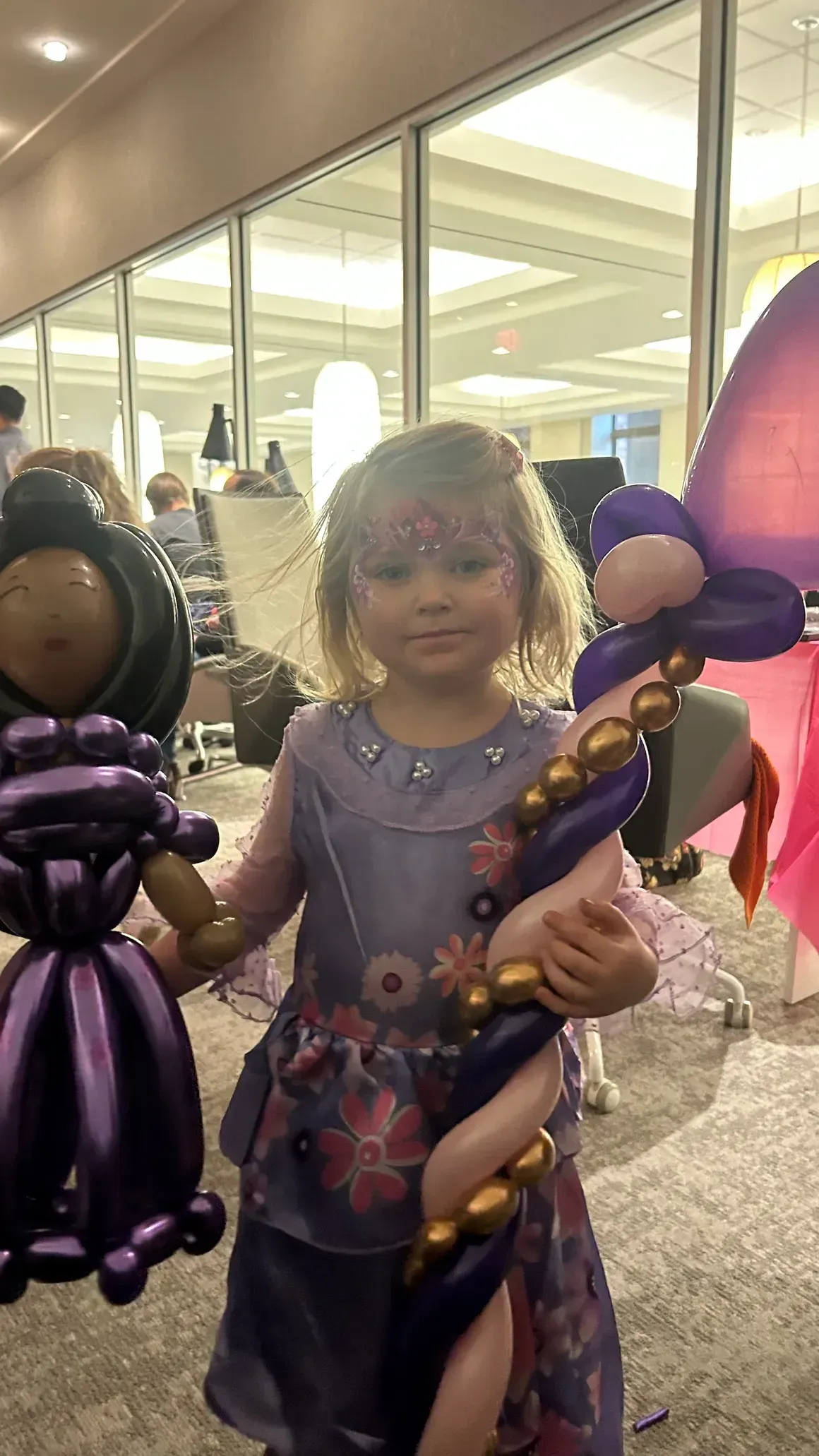 Girl in purple dress holds balloon dolls. Indoor event with large windows.