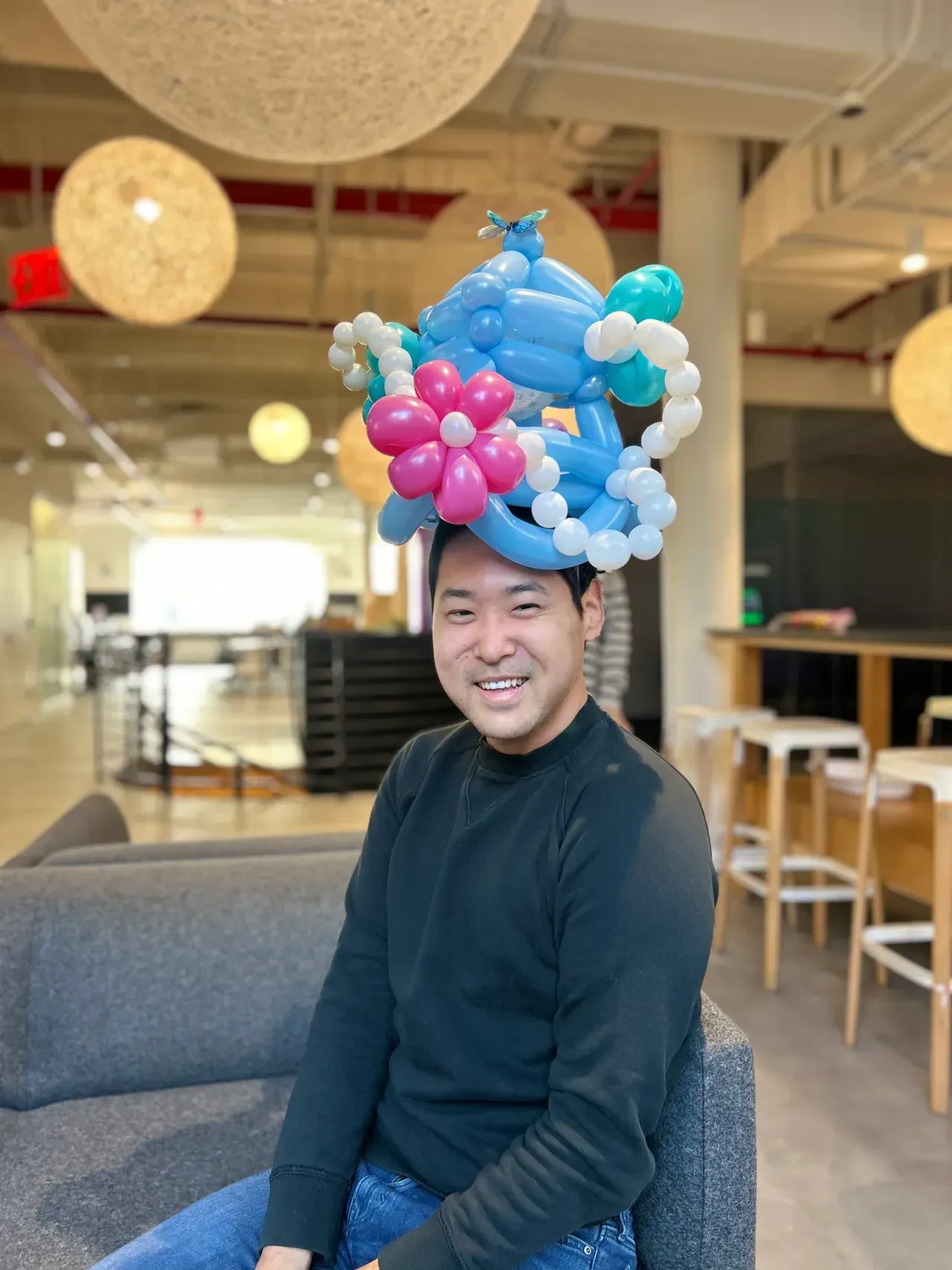 Man smiling, wearing a blue, pink, and white balloon hat, sitting on a gray couch in an office.