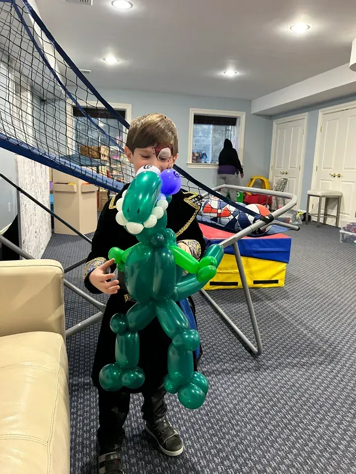 Boy holding a green balloon dinosaur with a Santa hat. Indoors with a volleyball net.