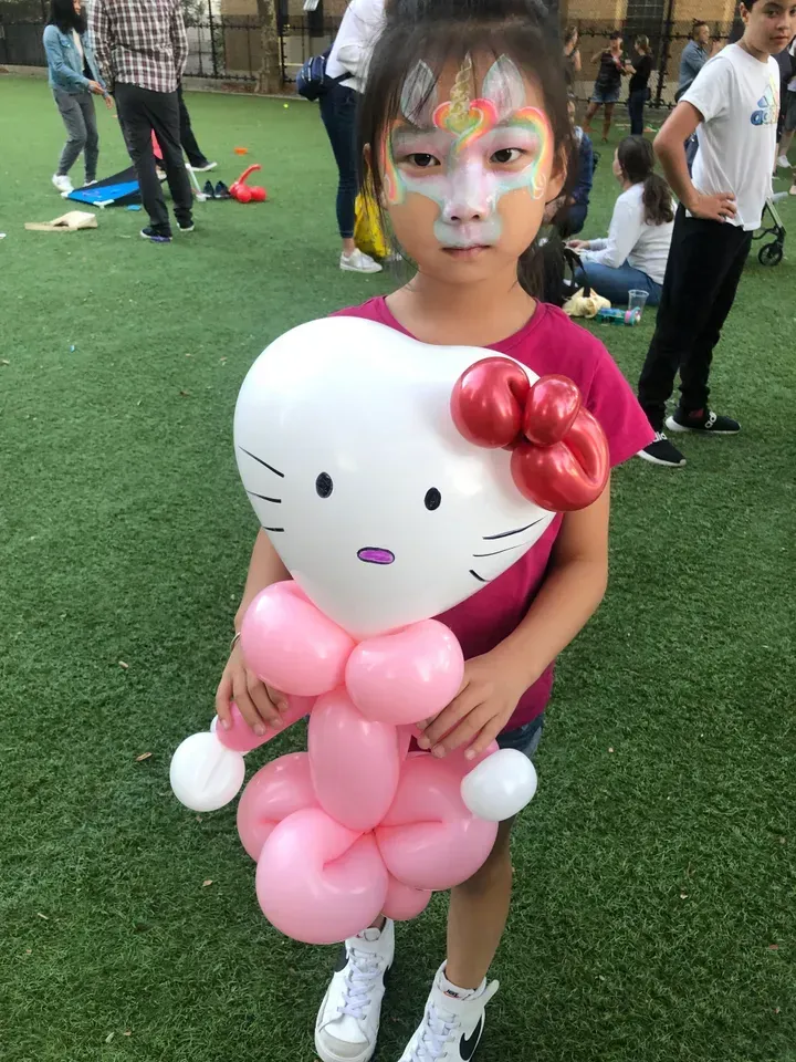 Girl with face paint holding a pink Hello Kitty balloon sculpture, outdoors on grass.