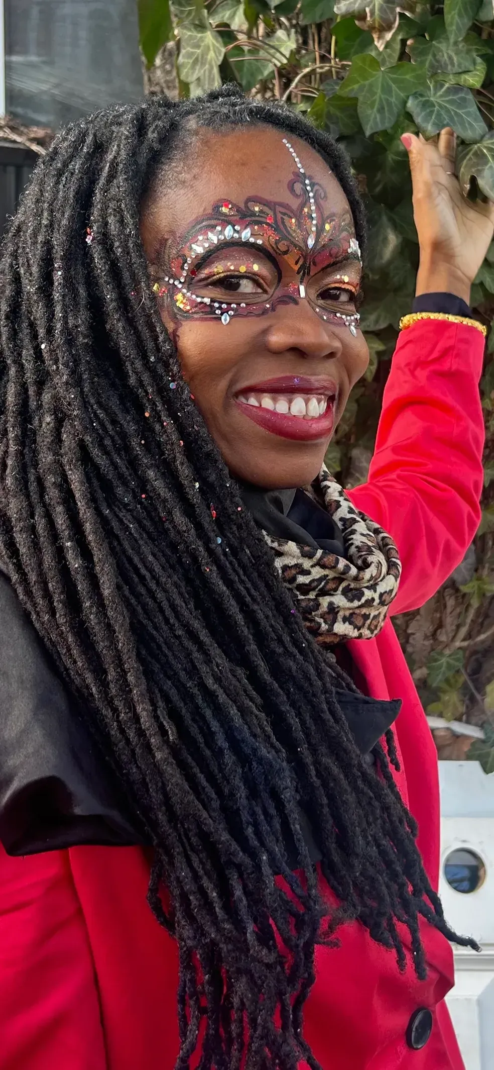 Woman with face paint and dreadlocks smiling, wearing a red coat, and reaching out toward greenery.
