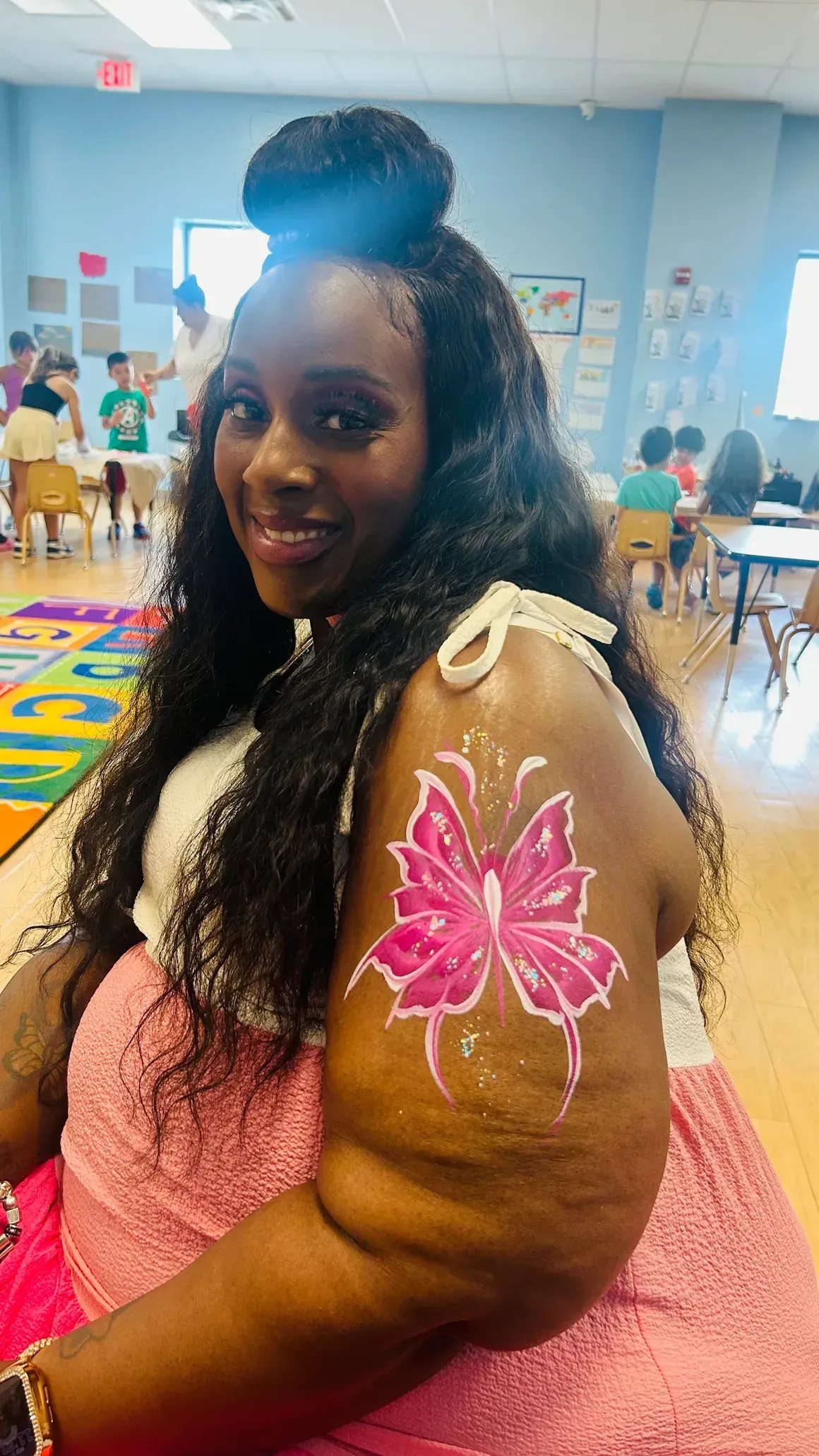 Woman with butterfly arm painting, smiling in a classroom with children.