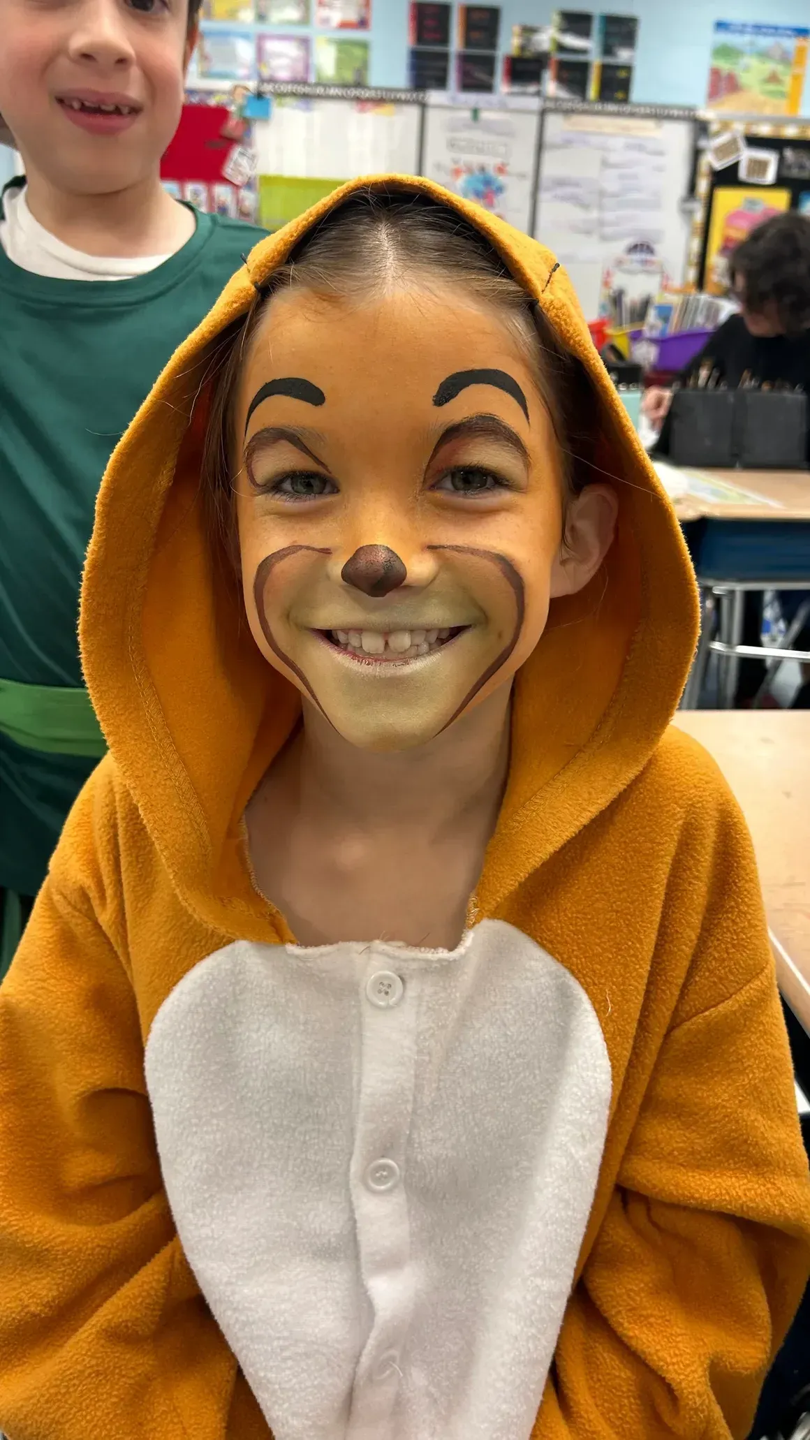 Child in Simba costume, face painted, smiles. Orange hooded jumpsuit, classroom setting.
