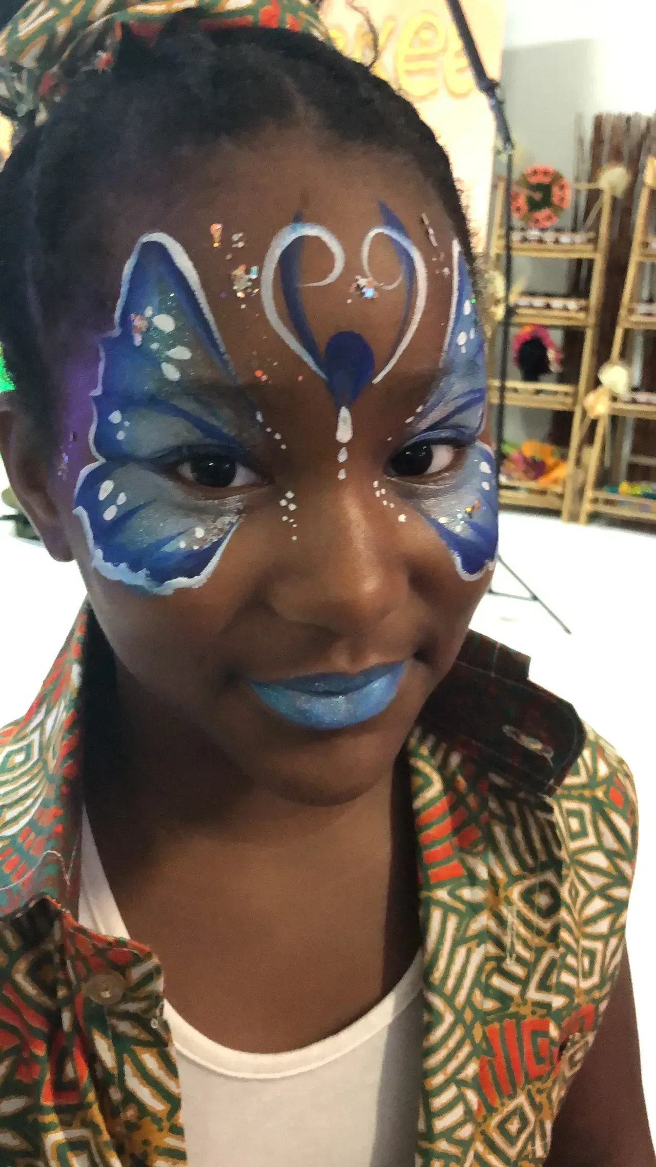 Girl with butterfly face paint, blue lipstick, and colorful patterned shirt smiles at the camera.