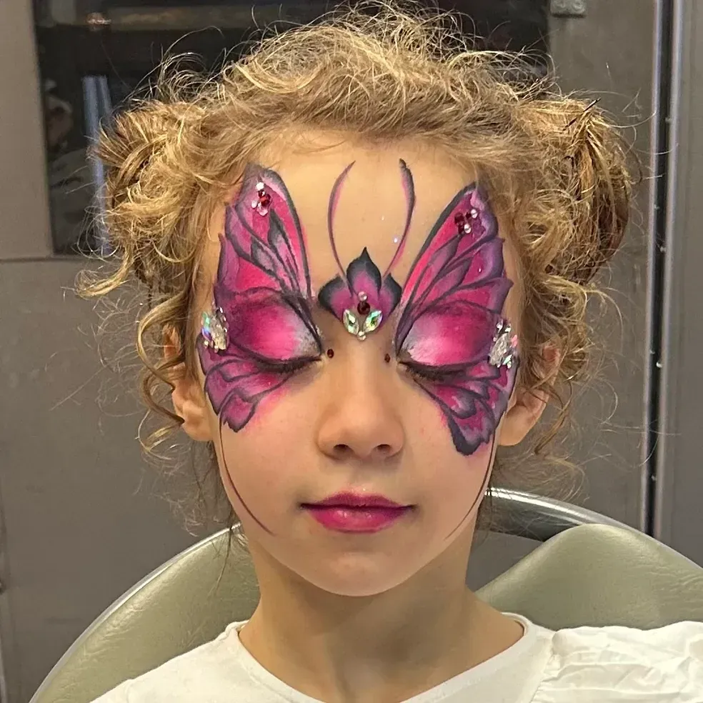 Girl with butterfly face paint, pink and jewel accents, eyes closed, smiling slightly.