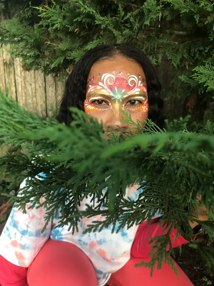 Person with face paint peeking through green foliage, wearing tie-dye and red.