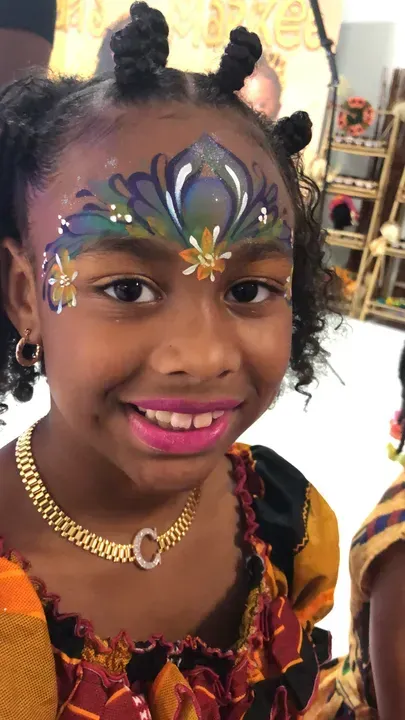 Girl with face paint and pink lipstick smiles, wearing a gold necklace, and a colorful patterned top.