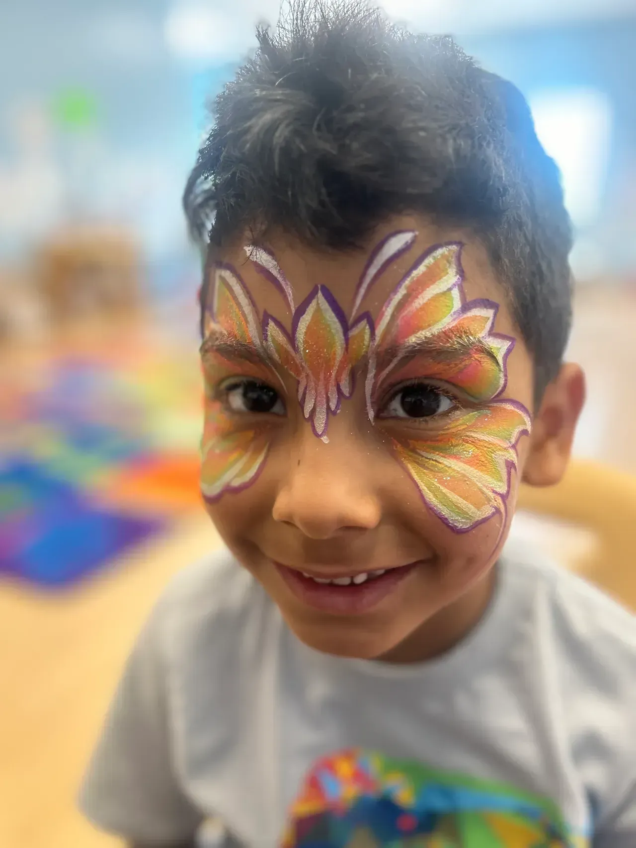 Boy with butterfly face paint smiles, indoors, blurry background.