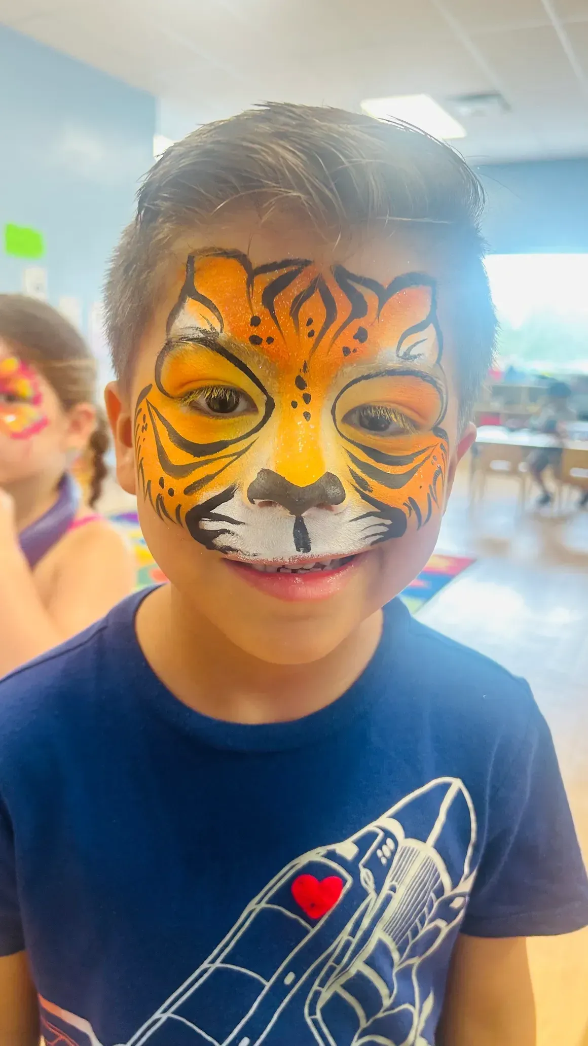 Boy with tiger face paint smiles, indoors, orange and black stripes.