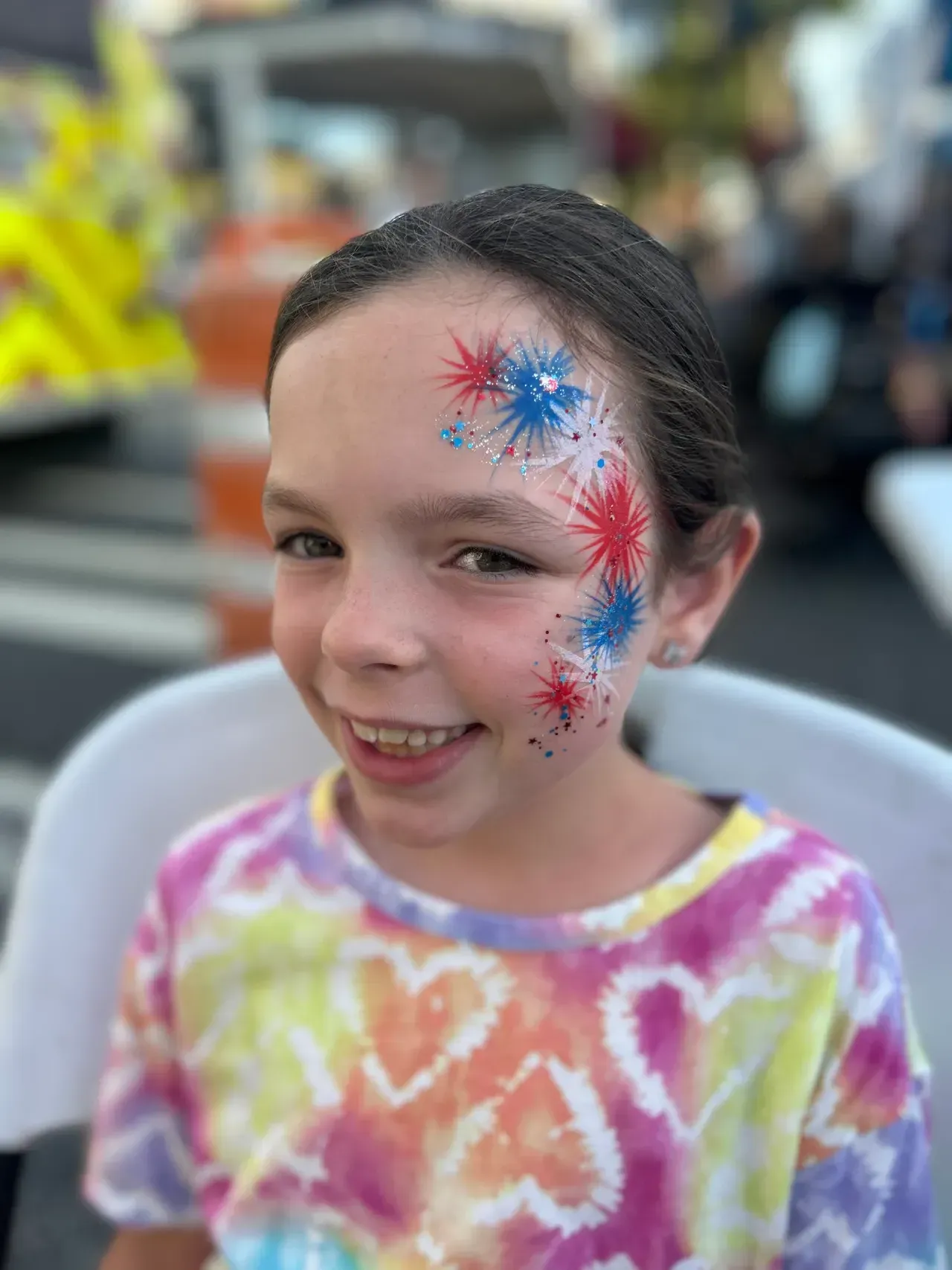 Girl with patriotic face paint smiles, wearing tie-dye shirt outdoors.