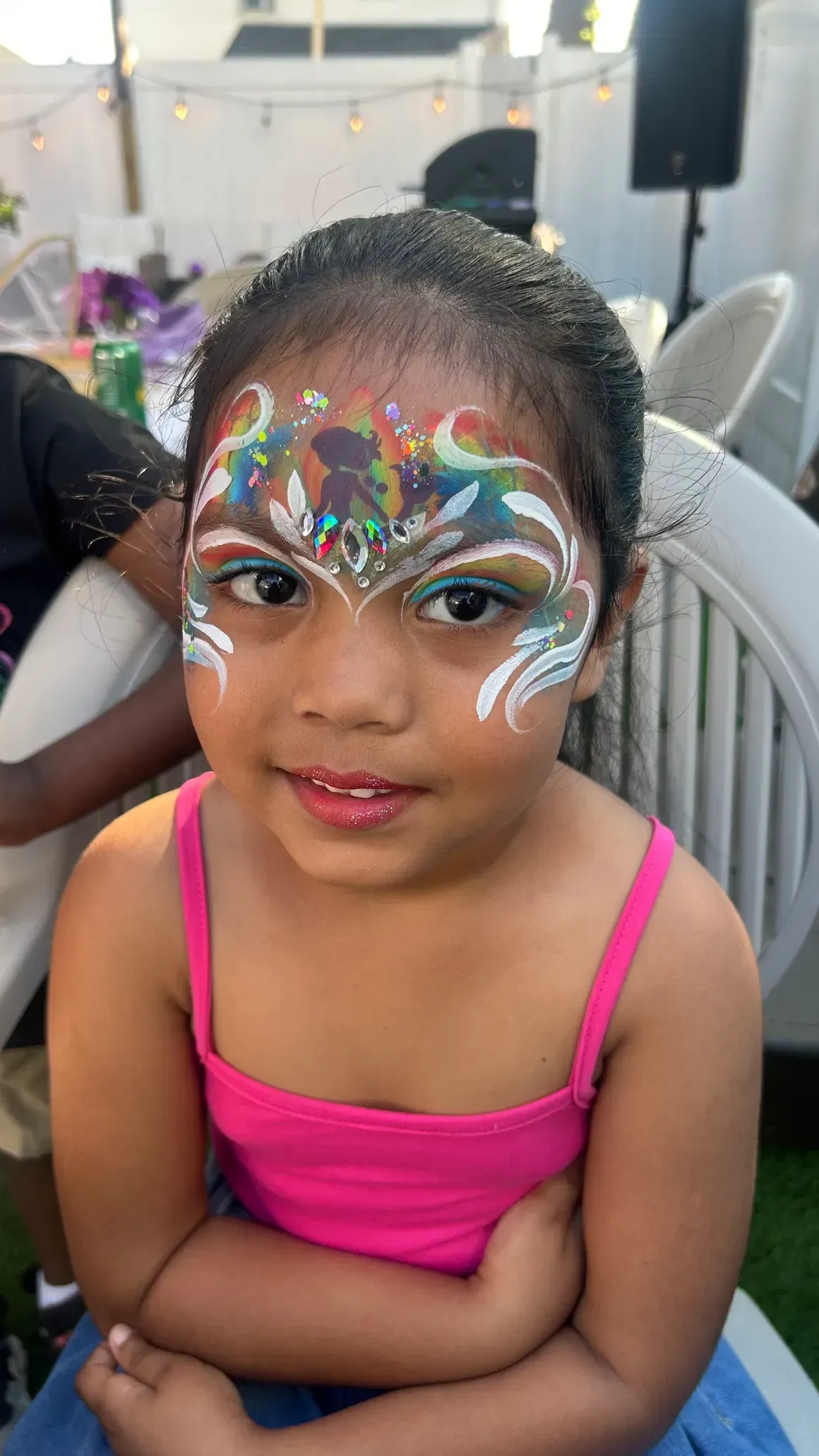 Girl with colorful face paint smiles, arms crossed, wearing a pink top, outdoors.