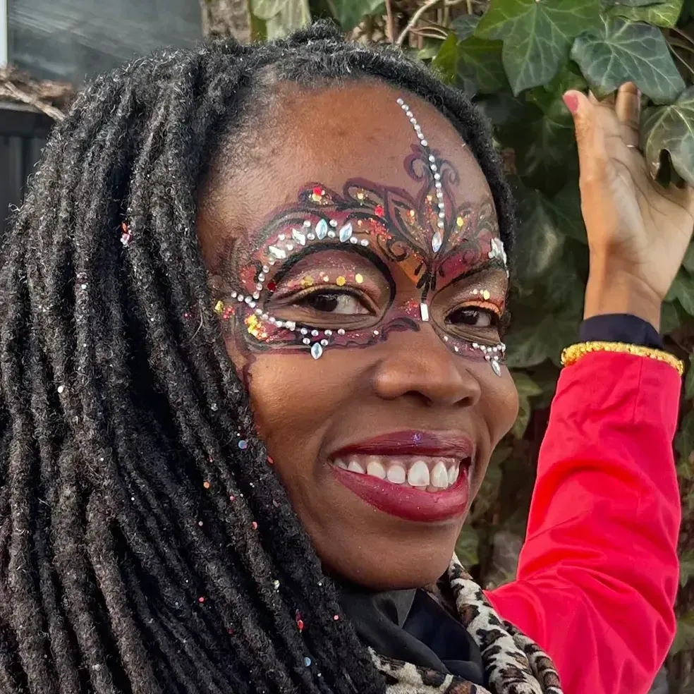 Person with face painted mask, smiling, reaching toward green leaves, red jacket.
