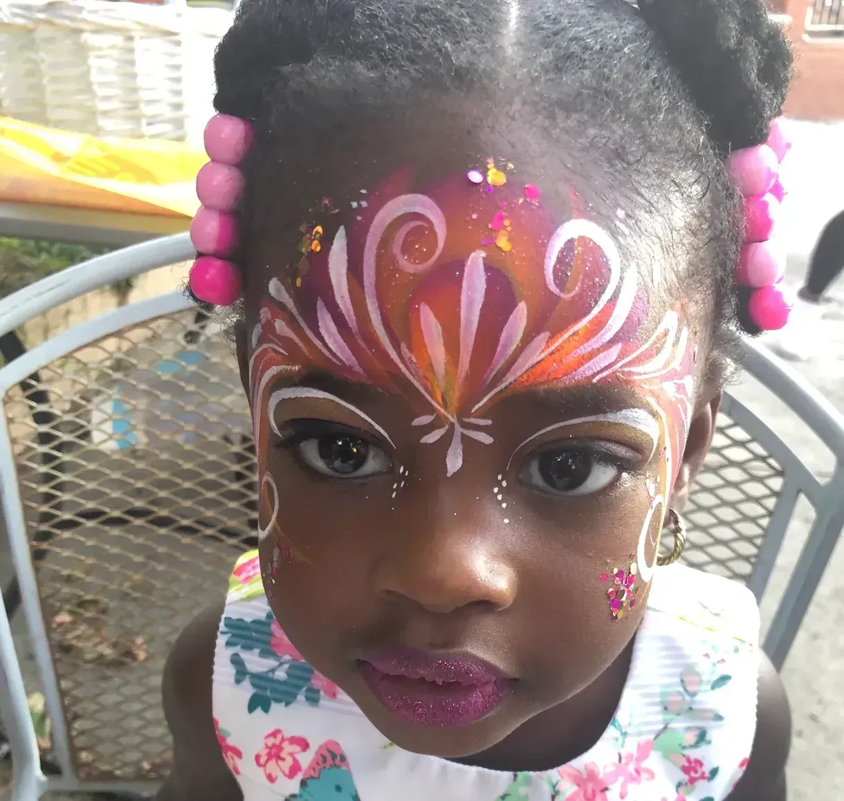 Girl with face paint design, outdoors. Eye-level shot. Face paint is white, pink, orange, glitter. Wearing floral top.