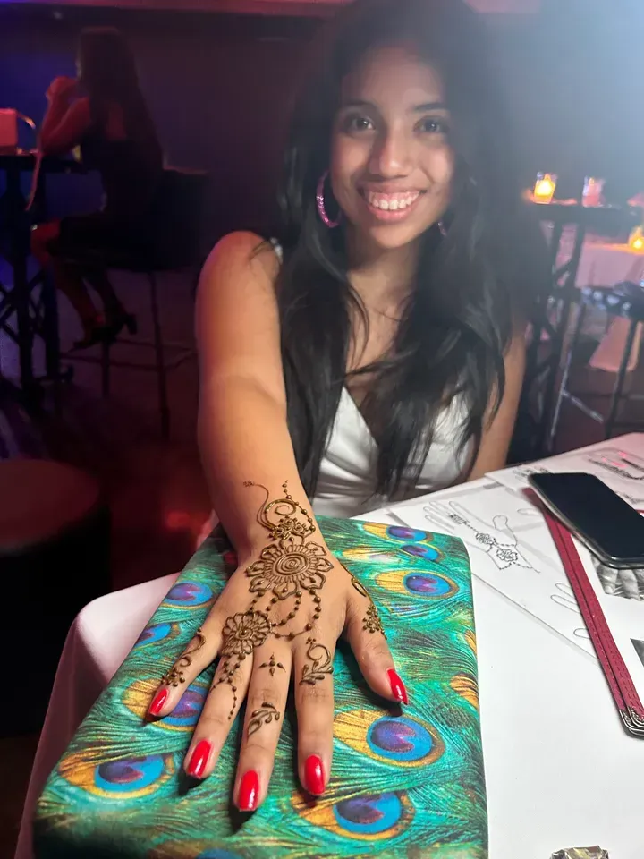 Woman with henna on hand, smiling. Red nails, peacock clutch, restaurant setting.