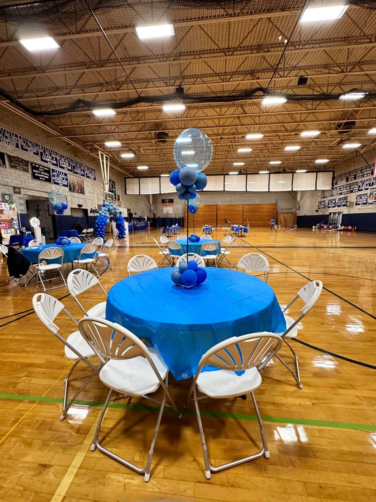 Round tables with blue tablecloths and white chairs in a gymnasium, decorated with blue balloons.