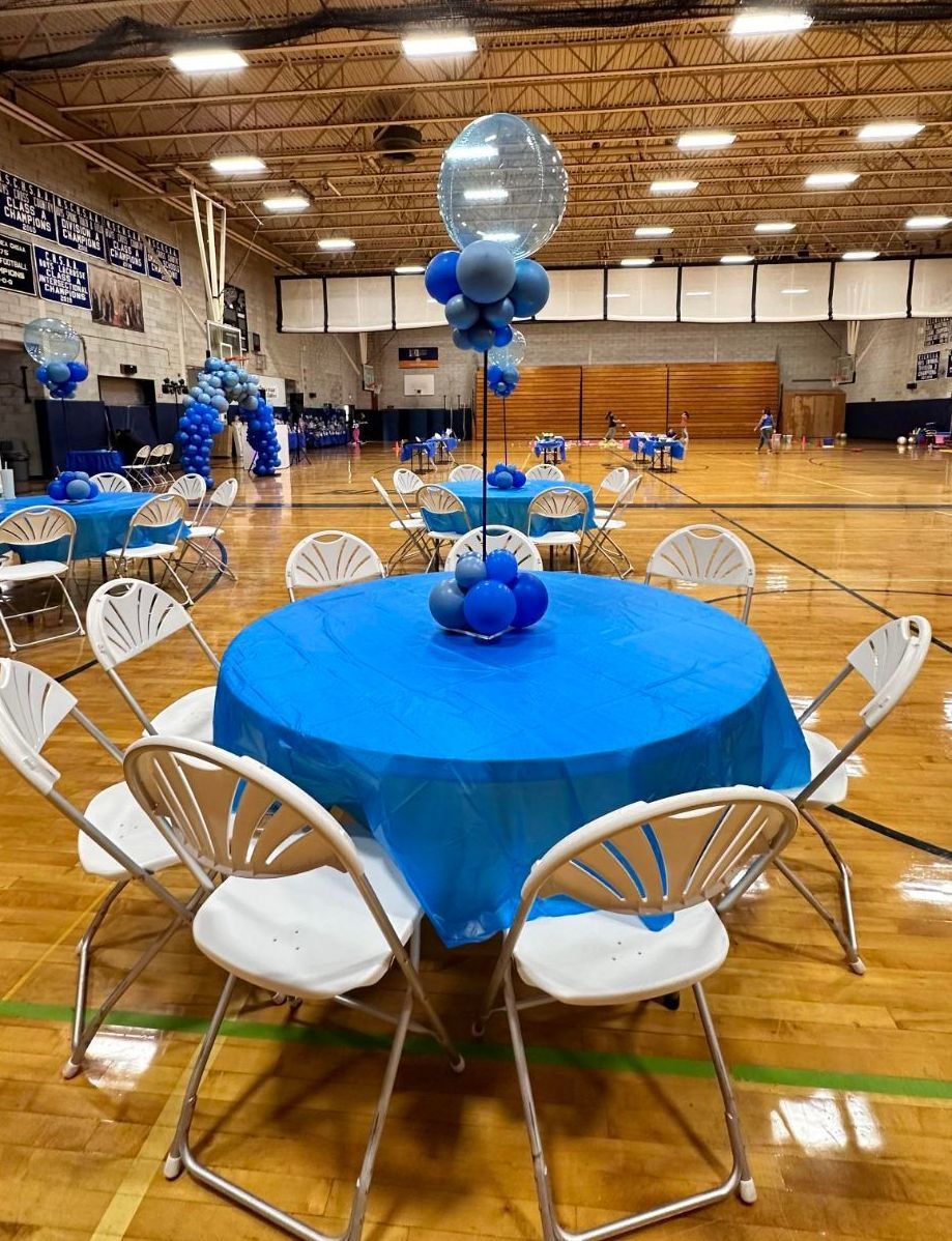 Round tables with blue tablecloths and balloon decorations in a gymnasium. White chairs surround the tables.