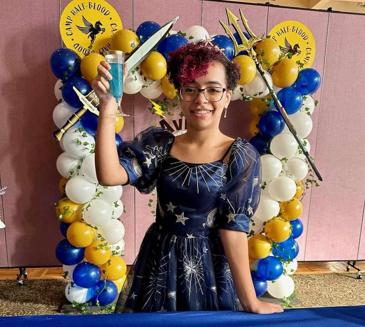 Young person with short curly hair, in starry dress, holds blue drink, smiles in front of balloon arch.