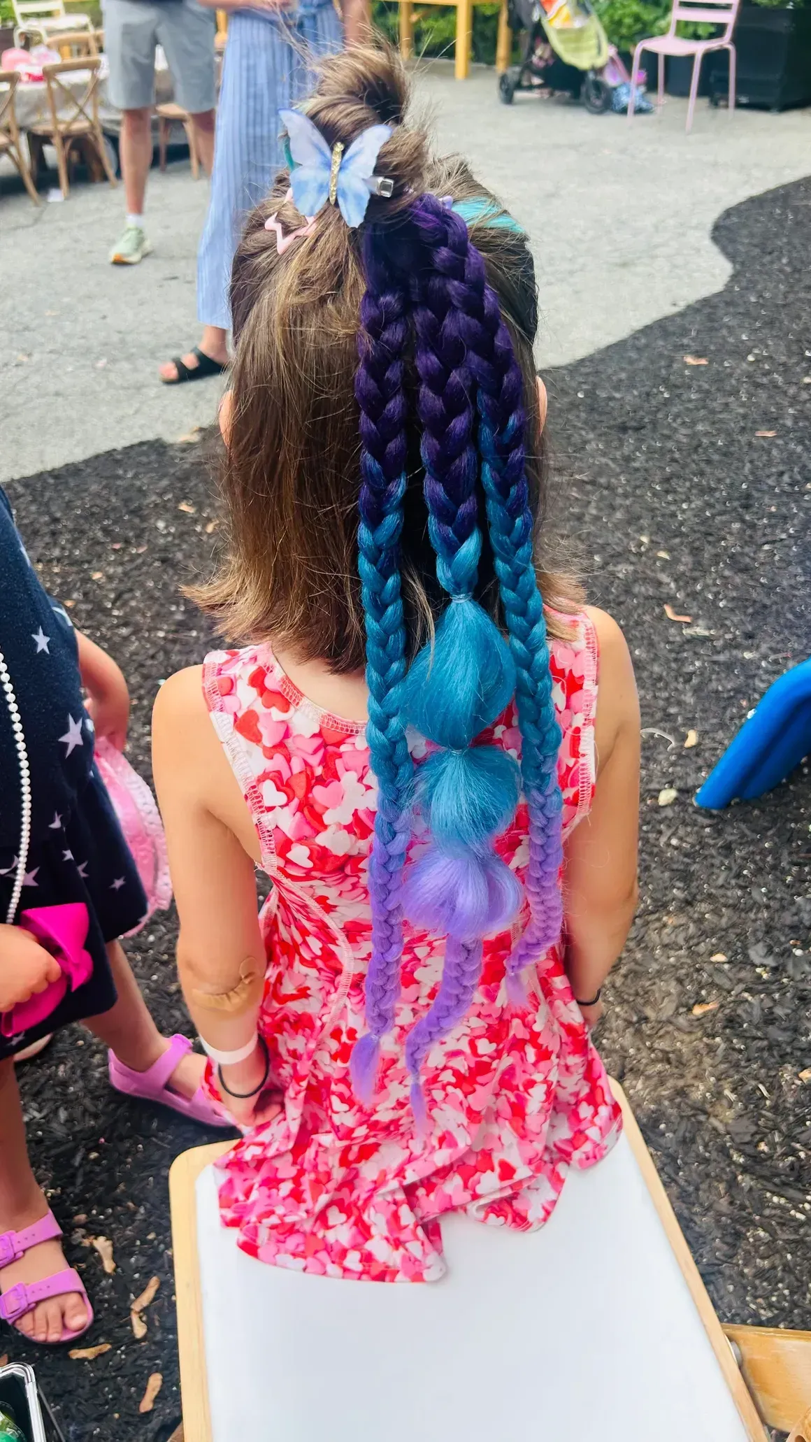 A little girl with purple and blue braids is sitting on a bench.