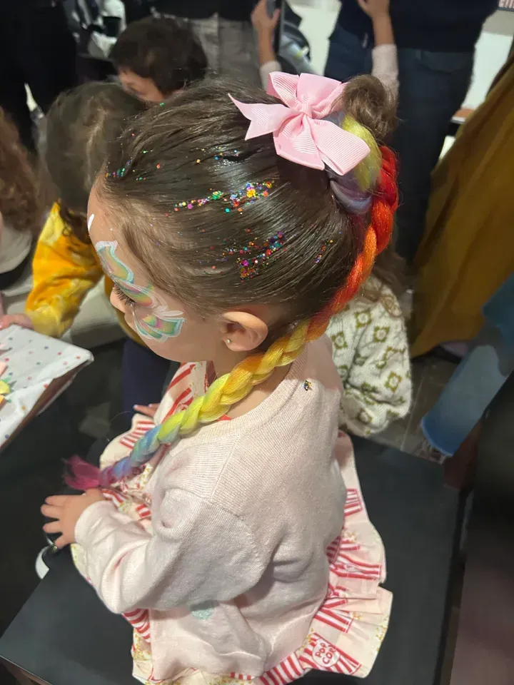 Girl with rainbow braided hair, pink bow, face paint, and glitter.