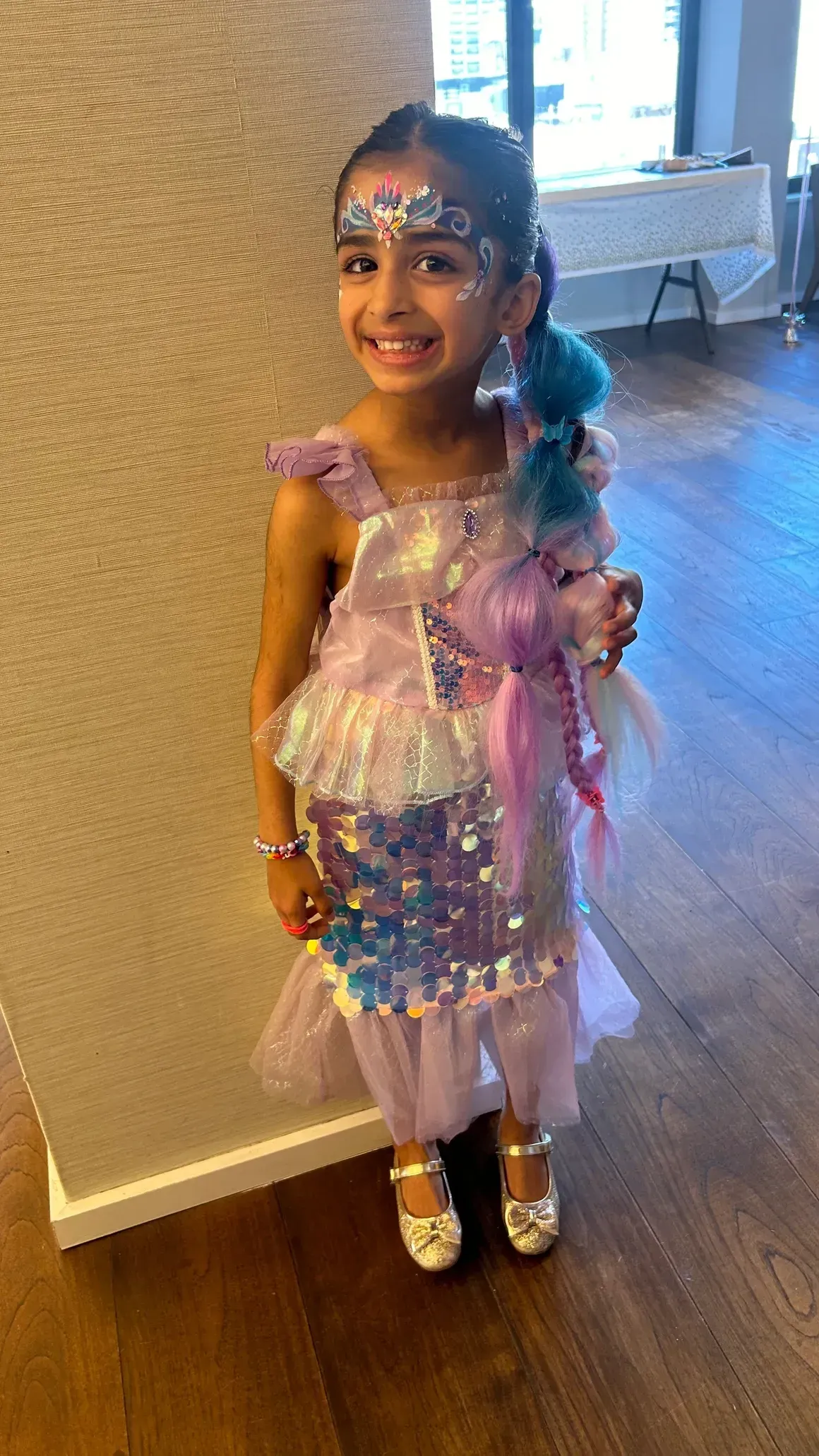 Girl in a sparkly mermaid costume, smiling, with face paint and blue hair, in a room.