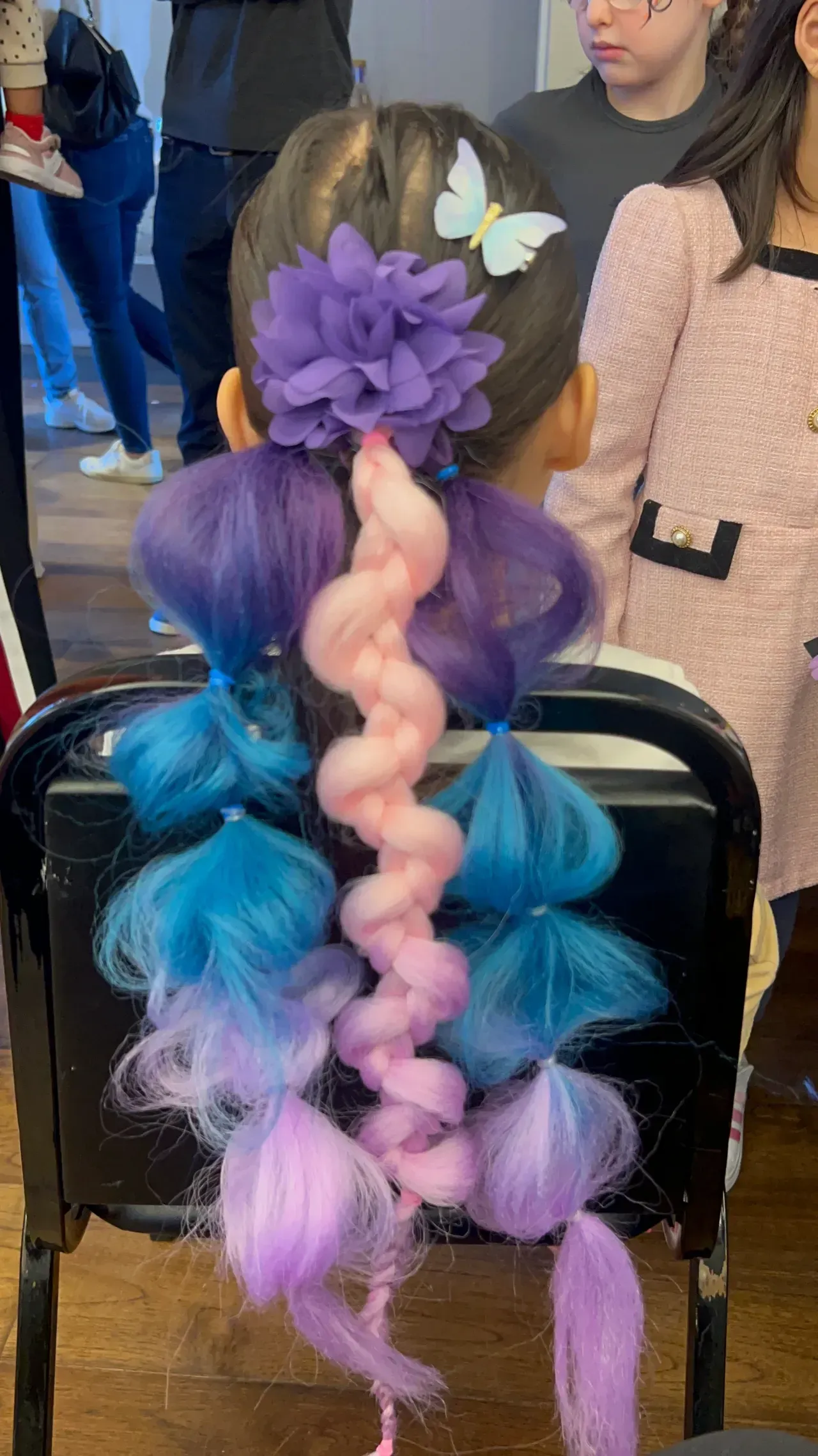 Child's hair styled with purple and blue sections, a pink braid, and a purple flower hair clip.