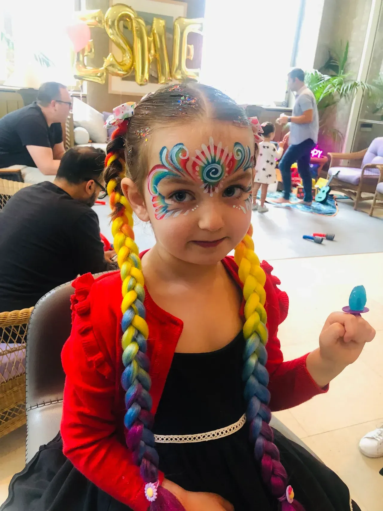 Girl with face paint and rainbow braids at a party, holding a ring pop, wearing a black dress and red jacket.