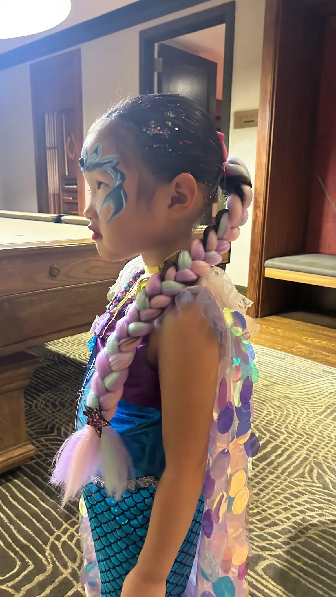Young child in mermaid costume with face paint, standing indoors near a pool table.