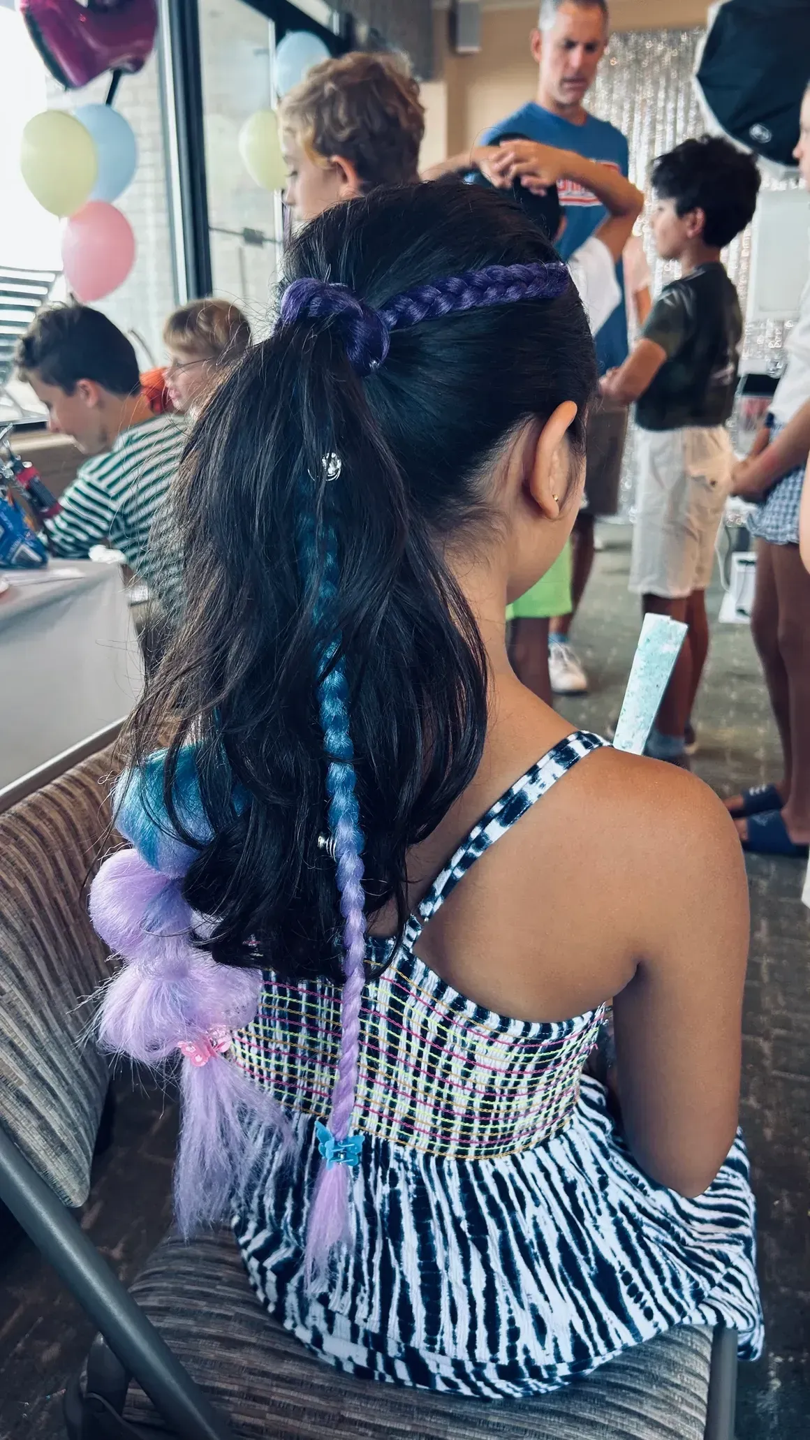 Girl with decorated hair, back to camera, at a party. Blue and purple accents, seated, with other people in background.