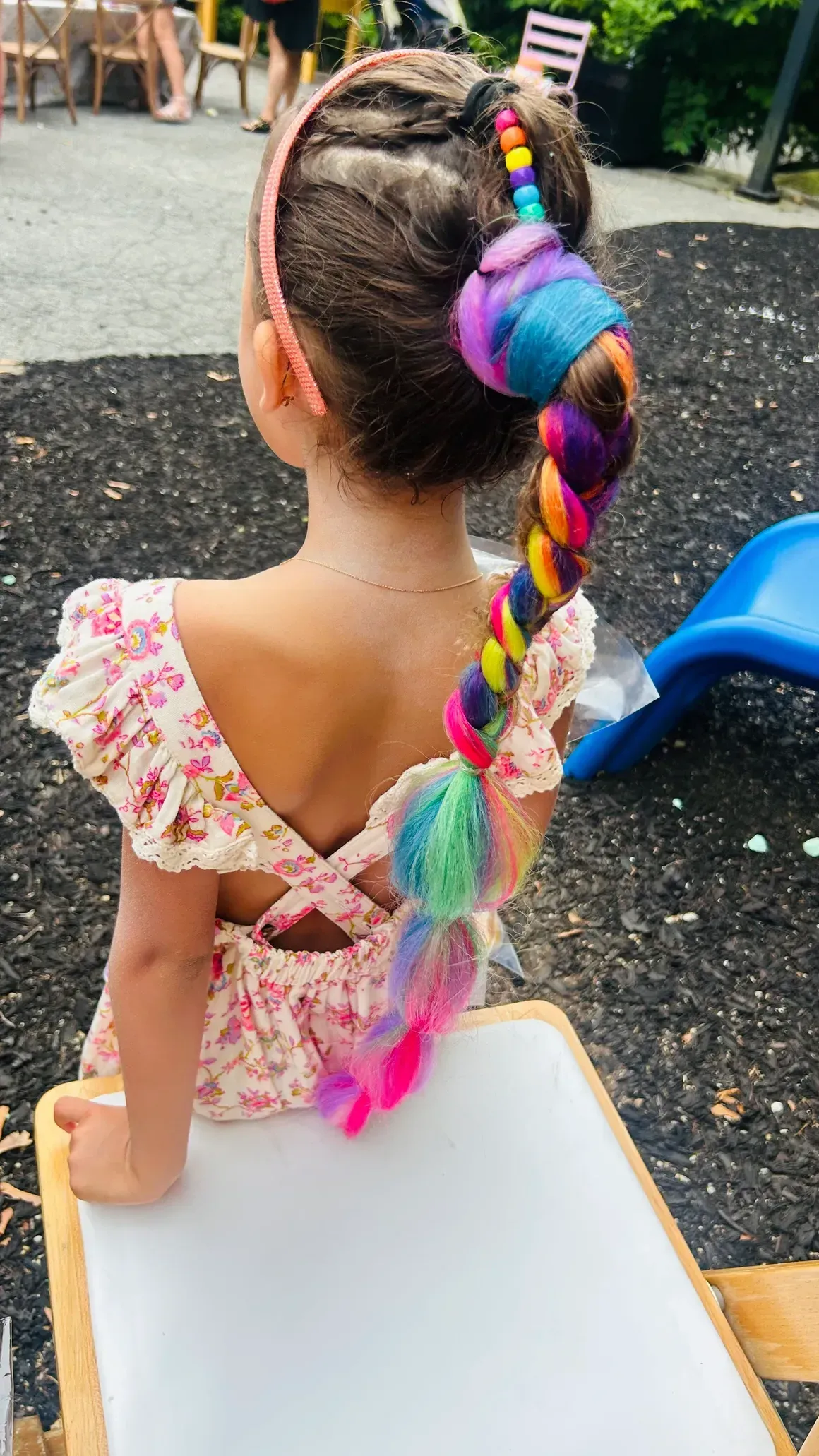 Girl with braided hair and colorful extensions, wearing a floral dress, outdoors.