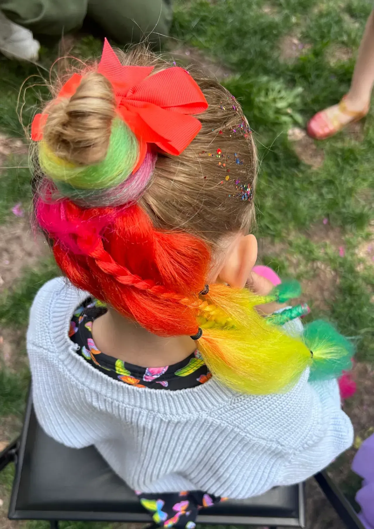 A little girl with a bun and rainbow hair is sitting on a chair.