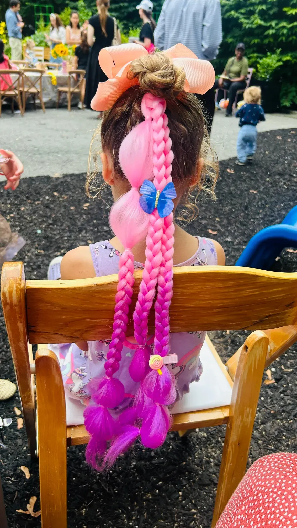 Girl with elaborate pink braided hairstyle seated outdoors.